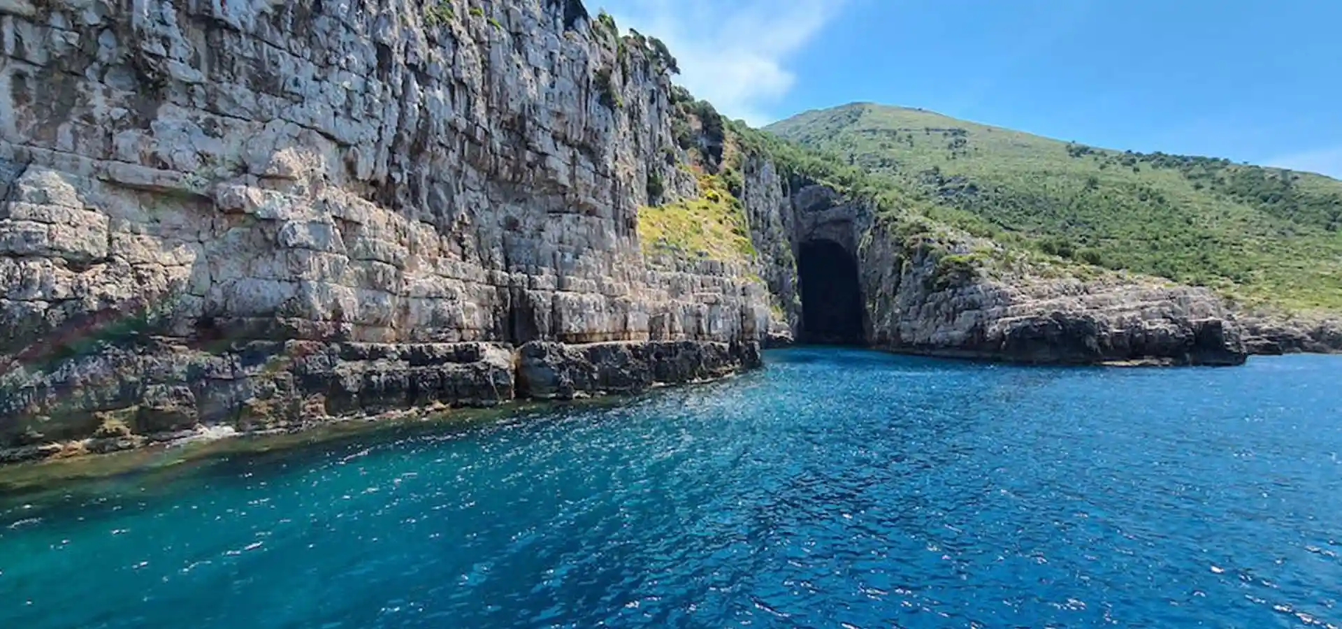Speedboat entering Haxhi Ali Cave with limestone arch over turquoise water, Karaburun Peninsula Albania