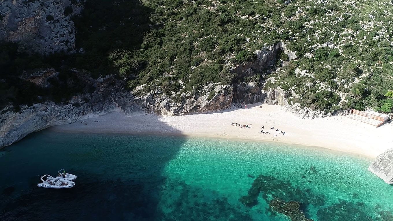 A stunning aerial view of Grama Bay, Albania, showcasing its secluded white-sand beach, crystal-clear turquoise waters, and dramatic rocky cliffs. A luxury speedboat floats nearby, offering an exclusive boat trip experience along the breathtaking Karaburun Peninsula. One of the top hidden gems on the Albanian Riviera, perfect for adventure seekers and nature lovers.